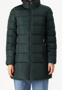 Mujer vestida con una chaqueta acolchada de color verde oscuro y con cuello alto, cremallera en el frente y bolsillos con cremallera, combinada con jeans azules.