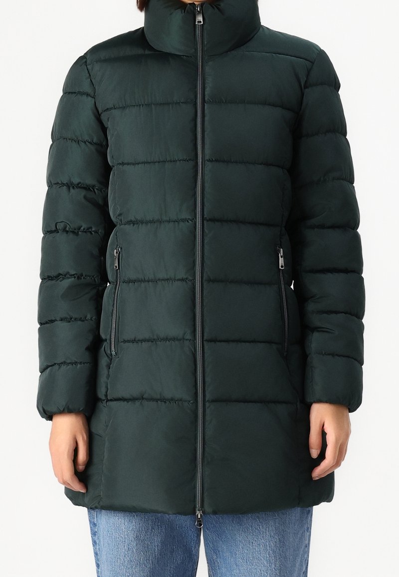Mujer vestida con una chaqueta acolchada de color verde oscuro y con cuello alto, cremallera en el frente y bolsillos con cremallera, combinada con jeans azules.