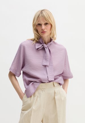 Blonde vrouw die een gestructureerde lavendelkleurige blouse met een strikceintuur draagt en crème kleurige, hoog- getailleerde, geplooide broek, handen in de zakken.