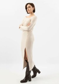 Robe beige à manches longues avec une fente sur le côté, silhouette ajustée, texture côtelée, associée à des bottes noires à talons hauts jusqu'aux genoux.
