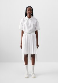 Hvit polo skjorte med mesh-detaljer, kombinert med en plisserte hvit skjørt og hvite joggesko. Merker seg spesielt en merket midjebånd.