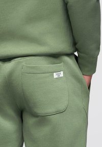 Personne portant un pantalon de survêtement vert olive et un sweat-shirt assorti, la main dans la poche latérale, poche arrière avec une petite étiquette blanche visible.