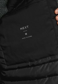 Juodas pūkinis švarkas su matinio paviršiaus interjeru. Yra audinio etiketė su užrašu "NEXT M" ir "MADE IN CHINA." Matomos siūlės.