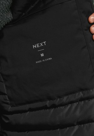 Juodas pūkinis švarkas su matinio paviršiaus interjeru. Yra audinio etiketė su užrašu "NEXT M" ir "MADE IN CHINA." Matomos siūlės.