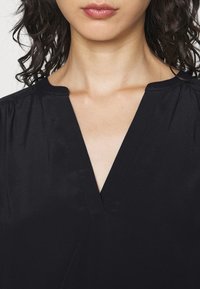 Top nero con scollatura a V, realizzato in tessuto morbido. Presenta maniche arricciate e una vestibilità morbida, mostrando un design pulito e minimalista.