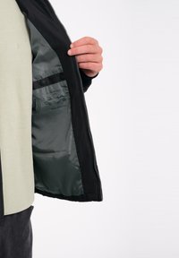 Chaqueta exterior negra con un forro de satén verde, que presenta una textura suave. Bolsillo interior con detalles impresos y una firma.