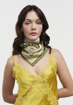Ženska z dolgimi temnimi lasmi, oblečena v rumeno majico z čipkastim robom in olivno zelen šal z vzorcem okoli vratu, na enobarvnem ozadju.