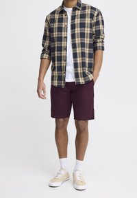 Camisa de franela a cuadros en azul marino y beige, camiseta blanca, pantalones cortos burdeos y zapatillas beige con suelas blancas. Conjunto casual con mangas enrolladas.