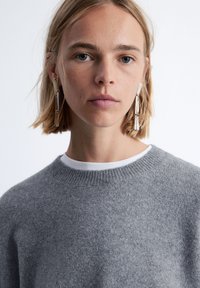 Grauer Wollpullover mit Rundhalsausschnitt, der eine sanfte Textur aufweist. Darunter ist ein weißes T-Shirt getragen, und lange, klare Ohrringe sind sichtbar.