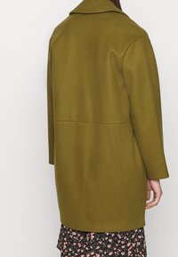 Kvinna i en lång olivgrön kappa över en svart blommig klänning, sedd bakifrån mot en enkel vit bakgrund.