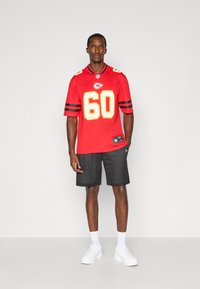 Sarkans Kansas City Chiefs kreklis ar dzelteniem numuriem, melnas svītras uz piedurknēm, apvienots ar melniem tīkla šortiem un baltām kedām.