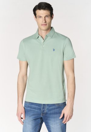 Uomo in piedi che indossa una polo verde chiaro e jeans blu su sfondo semplice.