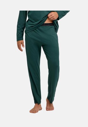 Uomo che indossa pantaloni lunghi da pigiama verde scuro e top, solleva leggermente la maglia per mostrare la fascia in vita, in piedi a piedi nudi su sfondo bianco.