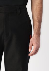 Sorte bukser med en glat tekstur og tilpasset pasform. Har en sidelomme og et lille rødt mærke på waistbandet.