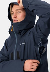 Giacca impermeabile blu navy con chiusure a zip, tasca laterale con zip, cappuccio regolabile e polsini in Velcro. Presenta il logo del brand sul petto.