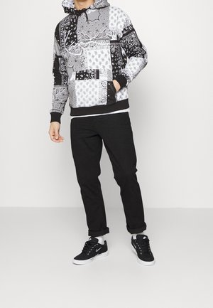 Hoodie in black and white bandana pattern with a drawstring hood, front pocket, and black ribbed cuffs. Paired with black pants and sneakers.