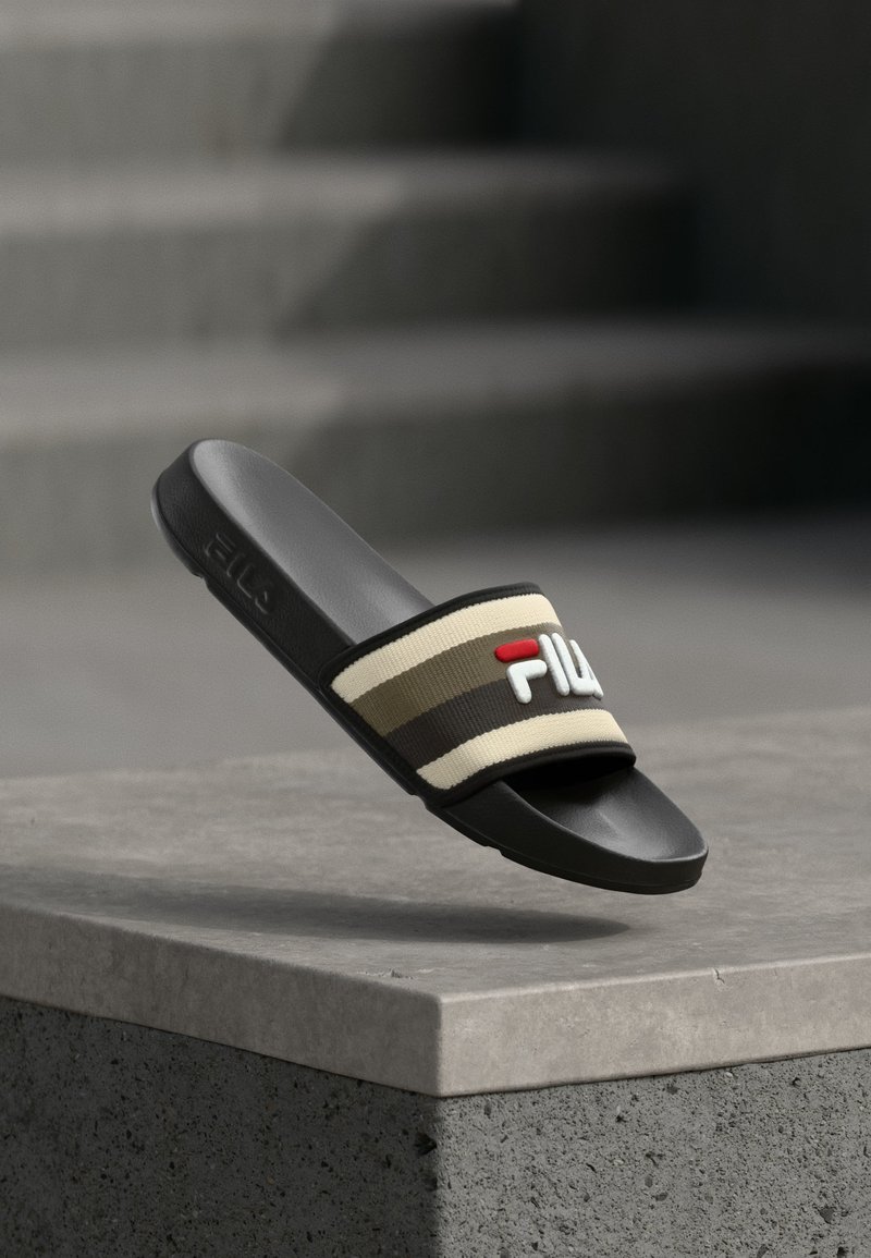 Μαύρο σανδάλι με σαγιονάρα και ριγέ μπεζ και καφέ υφασμάτινο λουράκι που φέρει το λογότυπο FILA, πάνω σε μια τσιμεντένια επιφάνεια.