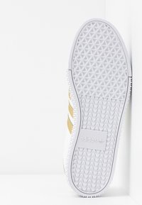 Suola di una sneaker bianca e dorata Adidas con schemi di grip testurizzati e logo visibile nella sezione centrale.