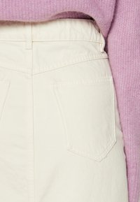 Pantalones de mezclilla blanca con una cintura alta y dos bolsillos traseros. La tela es gruesa con un acabado ligeramente texturizado. Combinados con un suéter de color púrpura claro.