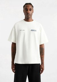 Camiseta blanca de algodón con mangas cortas, corte relajado, con texto y logotipo azules en el frente, y una sutil etiqueta en el dobladillo.