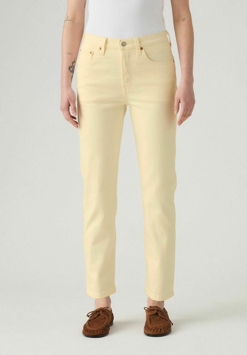 Personne portant un jean droit jaune clair, un débardeur blanc côtelé et des mocassins marron, debout devant un fond uni.