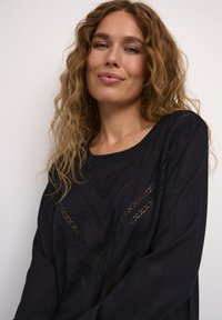 Blouse noire à manches longues avec des découpes intricates et une coupe décontractée. Texture lisse avec un col rond et des embellissements subtils.