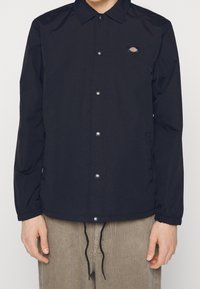 Man wearing a black button-up jacket with elastic cuffs and small logo on chest, paired with beige corduroy pants with visible drawstrings.