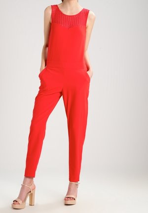 Kvinde iført en ærmeløs rød jumpsuit med blonder ved brystet, lommer og beige højhælede sandaler, stående mod en ensfarvet baggrund.