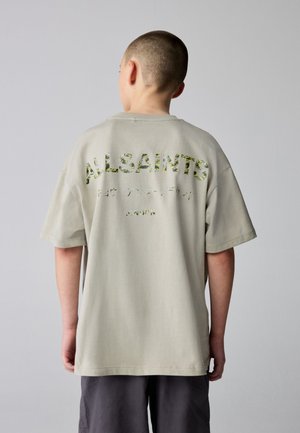 Person med kort hår iført en beige AllSaints oversized t-shirt med camouflageprint tekst på ryggen, parret med mørke shorts.