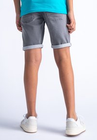 Šedé džíny šortky s ohrnutými lemy, mají zadní kapsy. Oblečené s bílými teniskami. Pozadí je jednoduché a bílé.