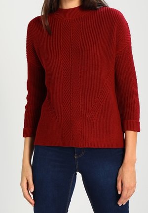 Pull en tricot côtelé rouge avec un col montant, des manches trois-quarts et des motifs texturés le long du corps. Associe à un jean foncé.