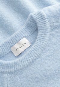 Svetlo modra pletena majica z rebrastim okroglim izrezom in belo tkanino z napisom "NOELLA", všito v notranji del ovratnika.
