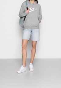 Grå huvtröja med vit logotyp, denimshorts och vita sneakers med rosa detaljer. Modellen bär en blå ryggsäck.