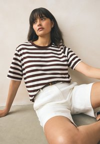 Barna és fehér csíkos póló kerek nyakkivágással és rövid ujjakkal, fehér magasított derekú rövidnadrággal párosítva. Sima szövet textúra.