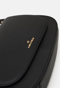 Borsa in pelle sintetica nera con una texture liscia, dotata di un cappuccio arrotondato e logo dorato in rilievo. Chiusura a cerniera lungo il bordo inferiore.
