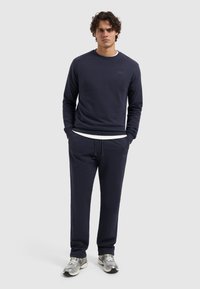 Marineblauwe sweatshirt en bijpassende sweatpants, gemaakt van zachte stof, met een klein logo op de borst. Model staat met handen in de zakken.