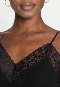 Svart linne med spetsdetaljer vid V-ringningen och axelbanden, samt en mjuk ribbad struktur på kroppen.