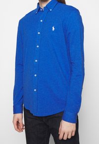 Niebieska koszula z długim rękawem, zapinana na guziki, wykonana z teksturowanej tkaniny, z klasycznym kołnierzykiem, białym logo i zapięciem na guziki.