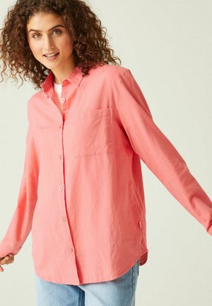 Giovane donna con capelli ricci che indossa una camicia corallo larga e dei jeans azzurri, sorridente con le braccia leggermente aperte.