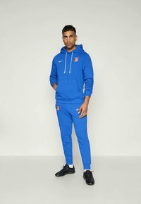 Sudadera azul y pantalones a juego hechos de una tela suave, con capucha, cordón y acentos de logo. Zapatillas deportivas negras completan el conjunto.
