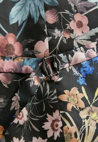 Tissu fleuri avec des fleurs roses, bleues et jaunes, doté d'une fermeture à boucle noire et argentée retenant deux sangles de tissu superposées.