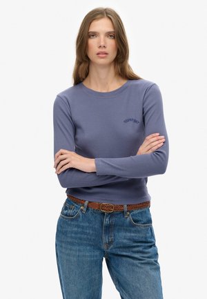 Joven mujer de cabello largo, con una blusa morada de manga larga y jeans azules, con los brazos cruzados, de pie frente a un fondo liso.