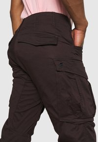 Mörkbruna cargo-byxor i mjukt tyg, med stora sidofickor och ett litet logopatch nära midjan.