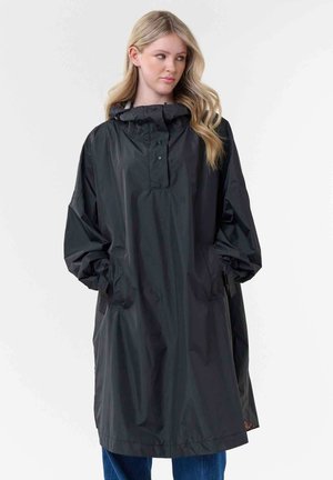 Blonde Frau mit lockerem schwarzen Regenmantel mit Kapuze, Hände in den Taschen, schaut seitlich vor schlichtem hellem Hintergrund.