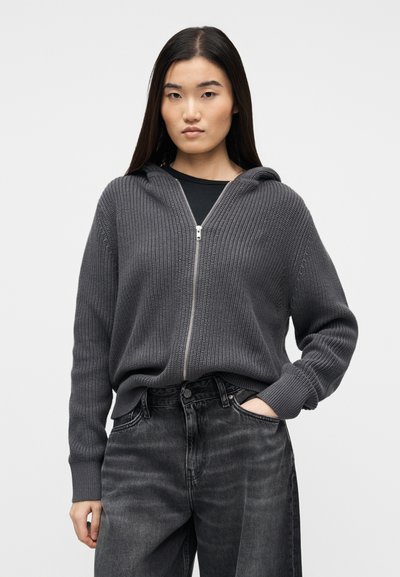 Νεαρή γυναίκα που φοράει σκούρο γκρι πλεκτό πουλόβερ με φερμουάρ και κουκούλα, και μαύρο ξεθωριασμένο τζιν, στέκεται με το ένα χέρι στην τσέπη της μπροστά σε απλό φόντο.