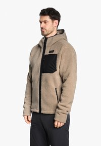 Veste en polaire beige avec une poche en nylon noire, fermeture éclair et capuche. Elle présente une texture douce et des poches latérales zippées.