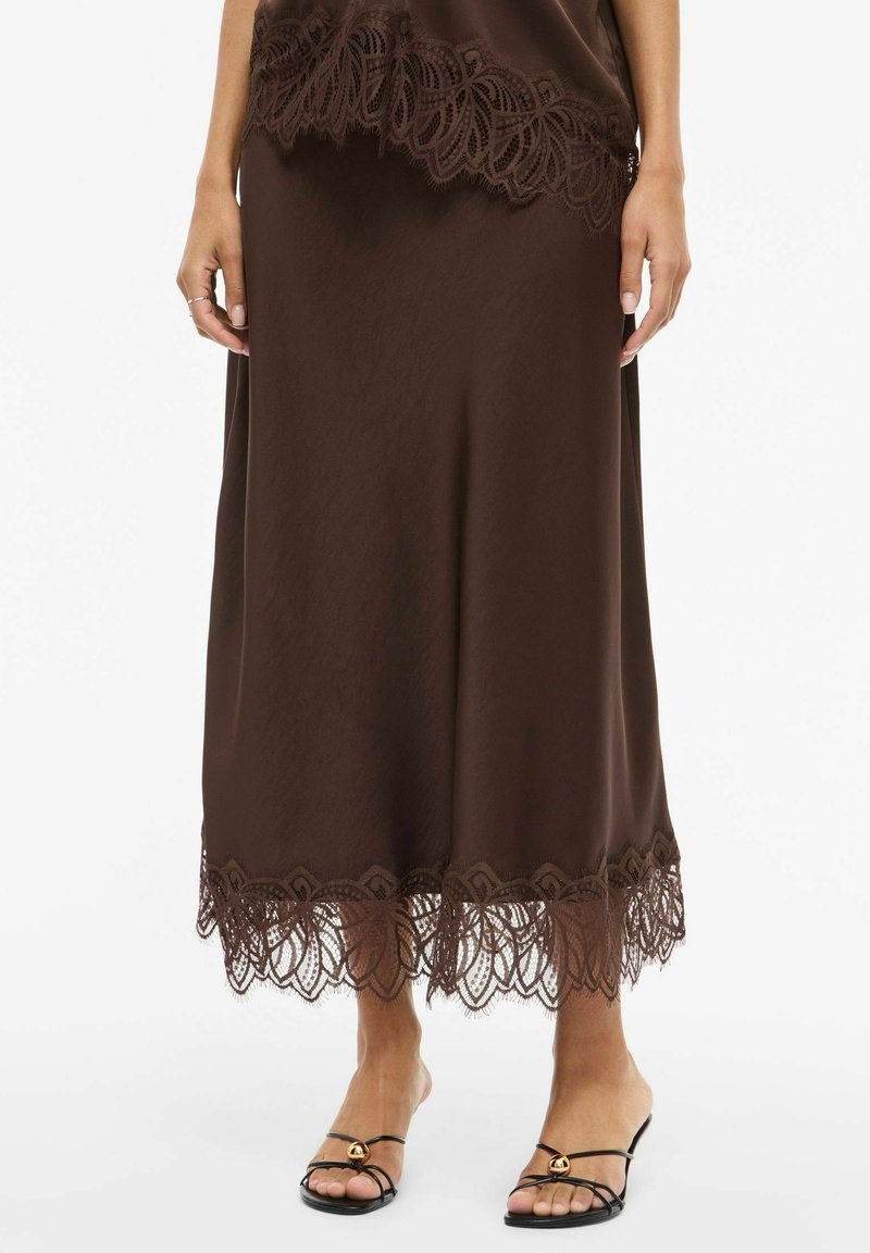 Brūna maksi svārki ar smalku mežģīņu apdari apakšmalā, nēsāti ar melnām siksniņu augstpapēžu sandales ar zelta rotājumiem.