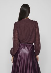 Blusa bordeaux con motivo a zig zag testurizzato e maniche a sbuffo, abbinata a una gonna plissettata di seta viola. Vista posteriore dell'outfit.