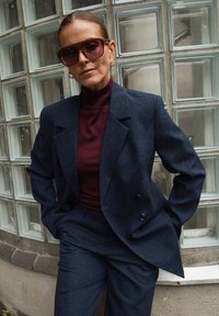 Ženska z velikimi sončnimi očali, mornarsko modrim dvokompletom in burgundskim puloverjem s kolikorom, stoji ob steni iz steklenih blokov.