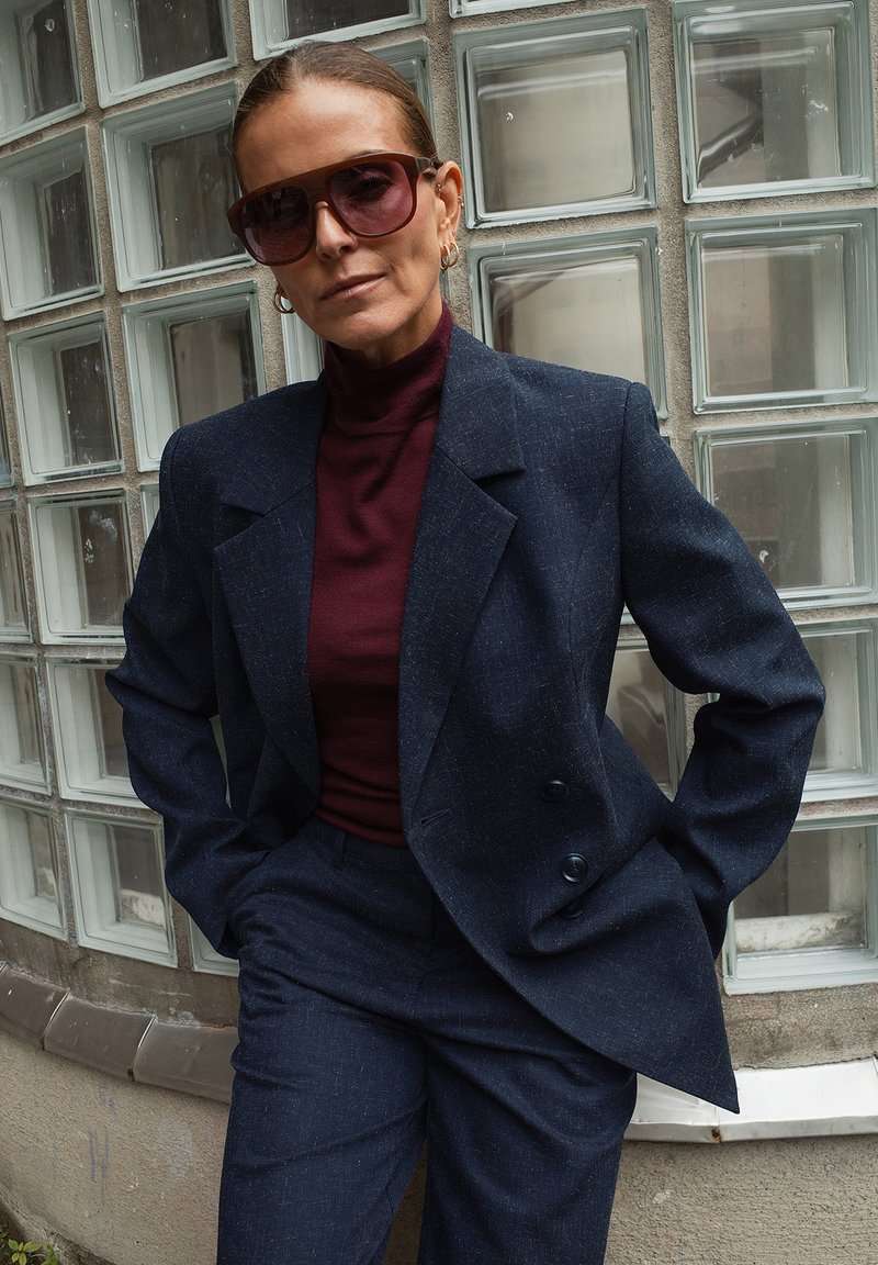 Ženska z velikimi sončnimi očali, mornarsko modrim dvokompletom in burgundskim puloverjem s kolikorom, stoji ob steni iz steklenih blokov.